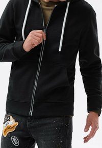 Personne portant un sweat à capuche noir zippé avec des cordons blancs et un jean noir orné de patchs colorés et de taches de peinture.