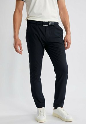 Homme portant un pantalon noir ajusté, une ceinture blanche, des baskets blanches et une chemise blanche à manches courtes sur un fond uni.