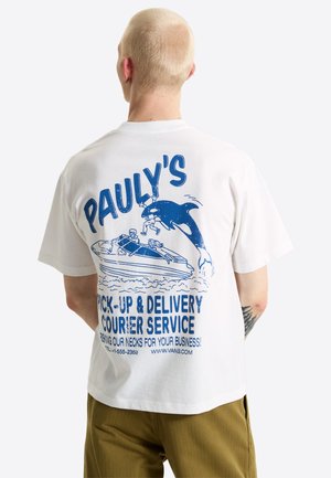 Person med kort blondt hår iført en hvid T-shirt med en blå grafik af en båd og en spækhugger med tekst om leveringstjeneste.