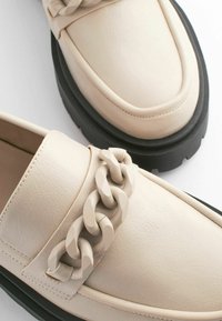 Beige nahkakengät paksulla mustalla pohjalla ja koristeellisella ketjuyksityiskohdalla edessä. Silkkinen pinta, jossa on vähäisesti ompelua.