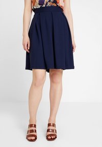 Gonna a pieghe blu navy con cintura aderente, realizzata in tessuto morbido e testurizzato. Abbinata a sandali marroni con cinturini e dettagli metallici.