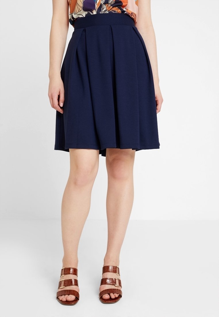 Gonna a pieghe blu navy con cintura aderente, realizzata in tessuto morbido e testurizzato. Abbinata a sandali marroni con cinturini e dettagli metallici.