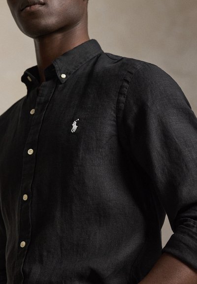 Άνδρας που φοράει μαύρο πουκάμισο με κουμπιά και λευκό κεντημένο λογότυπ ο παίκτη πόλο στο στήθος, με τα μανίκια ανασηκωμένα.