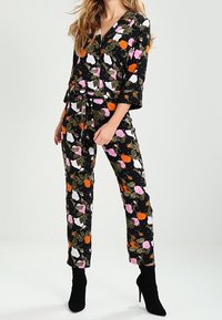 Svart blommig jumpsuit med livliga rosa, vita och orange blommor samt gröna blad, med V-halsringning, bälte och vida ärmar.
