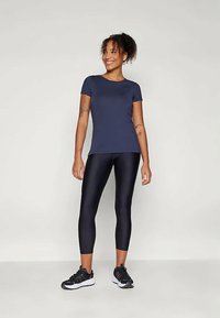 Námornícky modrý priliehavý krátky rukáv top s okrúhlym výstrihom, skombinovaný so lesklými čiernymi legínami po členky a čiernymi športovými topánkami, stojaci uvoľnene.