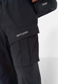 Zwarte cargobroek met meerdere zakken, voorzien van een gestructureerd uiterlijk en een zichtbare logo-detail aan de zijkant. Ontworpen voor functionaliteit en comfort.