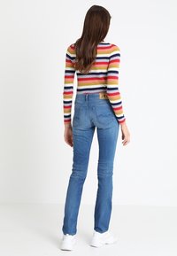 Vaqueros azules con una silueta ligeramente acampanada, que cuentan con bolsillos traseros y una etiqueta de cuero marrón en la cintura. Llevados con una camiseta de manga larga a rayas.