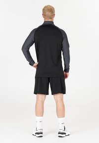 Ung man som står med händerna på höfterna, klädd i en svart långärmad tröja med mörka ärmar, svarta shorts, vita strumpor och vita sneakers.