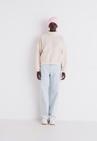 Helles beige Fleece-Half-Zip-Pullover, hellblaue gerade Jeans und eine rosa Strickmütze. Einfaches Design mit lässiger Passform.