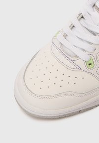 Κοντινό πλάνο της μύτης ενός λευκού αθλητικού παπουτσιού με τρύπες, λευκά κορδόνια και πράσινη λεπτομέρεια στο στοιχείο στερέωσης σε απλό ανοιχτό φόντο.