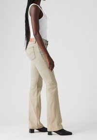 Pantalones vaqueros beige de campana con un lavado claro, con un diseño clásico de cinco bolsillos y una etiqueta de marca de cuero en la parte trasera. Botas negras de punta afilada usadas.