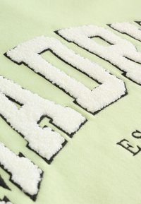 Tela verde claro con texto bordado en blanco y contornos negros, que presenta un dise�ño texturizado. No se ven detalles adicionales.