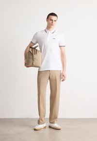 Hvit polo skjorte med svarte detaljer, beige bukser, holder en beige bag med håndtak og med stripete aksent. Hvite sneakers fullfører stilen.