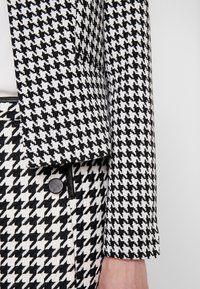 Blazer a motivo houndstooth in bianco e nero con un design leggermente strutturato, abbinato a una gonna coordinata e dettagli in bottone neri.