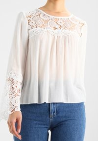 Blouse blanche à manches longues avec un empiècement en dentelle texturée, un corps plissé et des accents de dentelle sur les manches, portée avec un jean bleu.