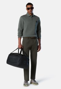 Maglione polo verde con tasca sul petto, pantaloni khaki, scarpe grigie e borsa a tracolla nera con logo "NORTH SAILS", portata da un uomo.