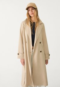 Manteau beige avec un design croisé, grand col et accents de boutons. Associé à une casquette assortie et un haut ajusté bleu marine.