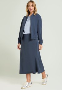 Granatowa kurtka bomberka i dopasowana spódnica midi z gładkiej tkaniny, zestawione ze srebrnymi trampkami. Projekt charakteryzuje się dopasowanym krojem i minimalistycznym stylem.