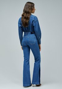 Camisa de denim y jeans acampanados en un lavado azul medio. Presenta bolsillos, cuello clásico y costuras visibles. Textura suave, ajuste entallado.
