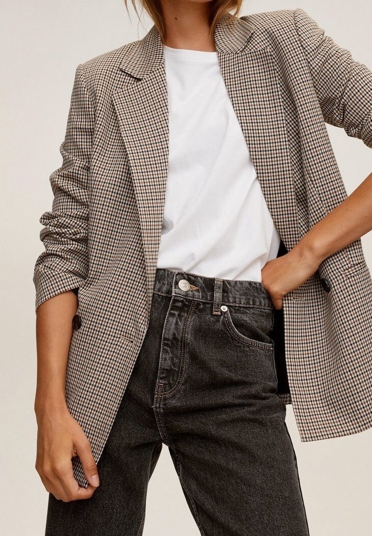 Femme portant un blazer beige et noir à carreaux sur un t-shirt blanc, associé à un jean en denim noir taille haute, main dans la poche.
