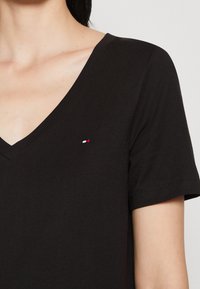 Sort bomulds T-shirt med V-udskæring, korte ærmer og et lille rødt, hvidt og blåt broderet logo på brystet. Glat overflade.
