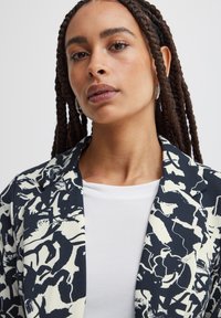 Patterned blazer in navy and cream, featuring abstract floral designs. Smooth fabric with a tailored fit and sharp lapels, layered over a white top.