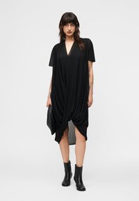 Γυναίκα που φοράει χαλαρό μαύρο φόρεμα με πτυχώσεις, μαύρα μποτάκια μέχρι τον αστράγαλο, κρατώντας μαύρο clutch, στέκεται μπροστά σε απλό φωτεινό φόντο.