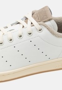 Bílá kožená teniska s perforovanými detaily, krémovými tkaničkami a béžovými akcenty. Má texturovaný hnědý jazyk a gumovou podrážku.