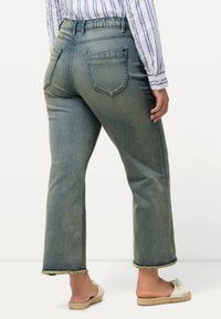 Jeans de mezclilla azul claro con corte recto, dobladillo deshilachado, cuatro bolsillos y cinturilla elástica, mostrados desde la parte trasera. Acabado texturizado.