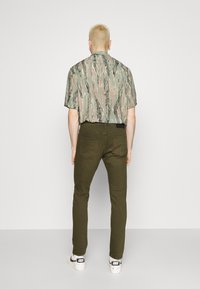 Camisa de manga corta en un patrón de camuflaje verde y rosa, combinada con pantalones verde oliva y zapatillas blancas, vista desde atrás.