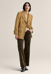 Beige kaksirivinen villatakki isoilla napeilla, kerma valkoinen pooloneule, ruskeat corduroy-housut ja mustat nilkkurit neutraalia taustaa vasten.