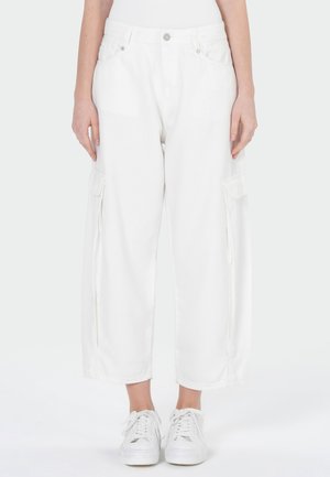 Persoon draagt witte wijde cargobroeken en witte sneakers, staand tegen een effen lichte achtergrond.