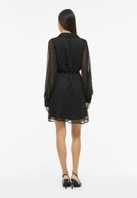 Czarna sukienka w białe groszki z kołnierzykiem, długimi przezroczystymi rękawami, taliowaniem i rozkloszowanym dołem. Gładka faktura i długość do kolan.