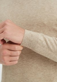 Pull en tricot beige avec des poignets côtelés. Le tissu est texturé avec un motif légèrement marbré, offrant une apparence lisse et confortable.