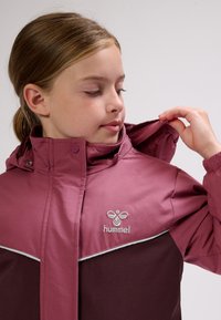 Ung person, der justerer huen på en tohjulet vinrød Hummel-jakke med høj krave og knappedetaljer.