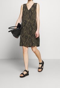 Klänning med V-ringning och ärmlös design, med en brun bas och svarta geometriska mönster. Tillsammans med svarta sandaler och en svart tote-väska.