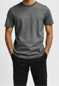 Man som bär en enkel mörkgrå t-shirt med kort ärm och svarta byxor, med händerna i fickorna, stående mot en neutral bakgrund.