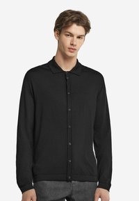 Svart långärmad cardigan med polokrage, framtill knappar och en mjuk textur, bärs över grå byxor.