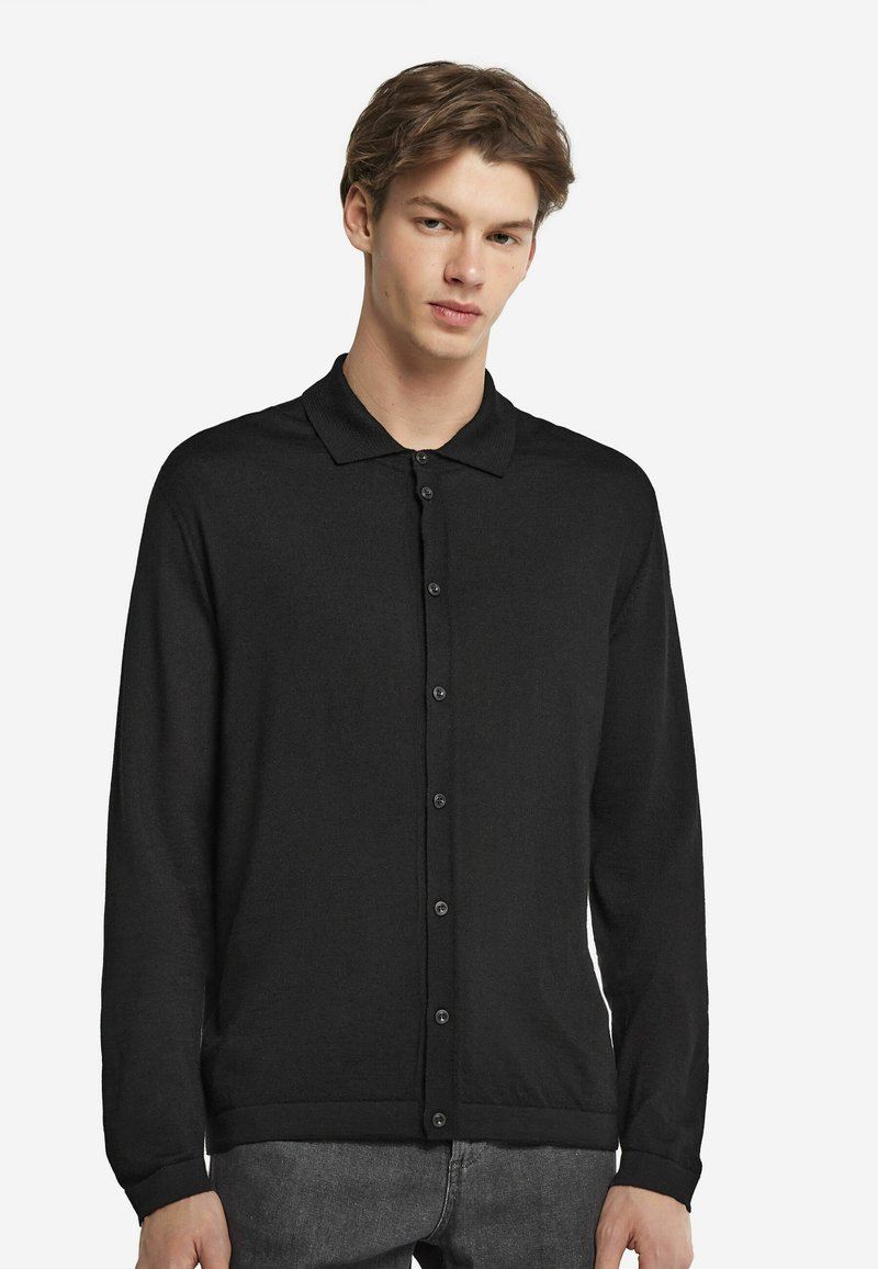 Svart långärmad cardigan med polokrage, framtill knappar och en mjuk textur, bärs över grå byxor.