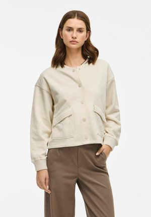 Light beige sweatshirt with a round neckline, snap buttons, two front pockets, and ribbed cuffs, paired with brown pants. Smooth texture.