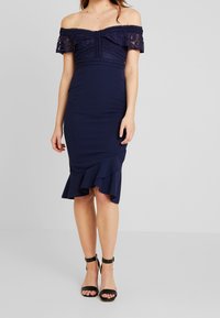 Navy avaxelklänning med spetsdetalj, figursydd bodice och volangkant. Tillverkad av ett mjukt, elastiskt tyg. Svarta högklackade sandaler.