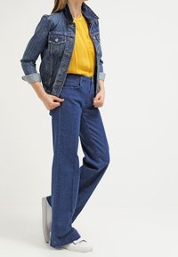 Giubbotto di jeans con taschini sul petto, blusa gialla e jeans blu a gamba larga. Sneakers bianche completano l'outfit. Denim azzurro, vestibilità casual.