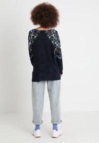 Persona con capelli ricci che indossa un maglione a fiori scuri, jeans chiari arrotolati, calzini viola e sneaker bianche Nike, in piedi di spalle.