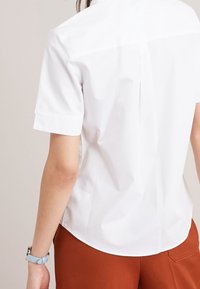 Weißes Kurzarmhemd mit glatter Textur und Rüschennaht auf dem Rücken, kombiniert mit braunen Hosen und einer hellblauen Uhr.