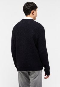 Granatowy sweter dzianinowy o ryflowanej strukturze, luźny krój i klasyczny okrągły dekolt, zestawiony z szarymi spodniami.