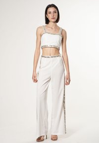 Top bianco corto e pantaloni a gamba larga con accenti argento su cinturini e cuciture laterali, texture liscia, silhouette aderente e sandali neutri.