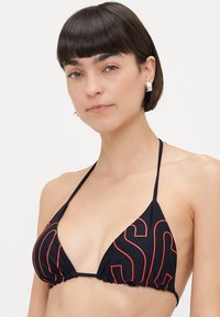 Kvinde med kort mørkt hår iført en sort halterneck bikini-top med røde omridser og sølvfarvede firkantede øreringe mod en hvid baggrund.