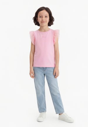 Ragazza con capelli corti e ricci, indossa una maglietta rosa con maniche arricciate, jeans azzurri e sneakers bianche, in piedi su uno sfondo bianco.