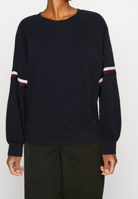 Felpa navy con maniche raglan, caratterizzata da strisce bianche e bordeaux sulle braccia. Tessuto morbido, vestibilità comoda e polsini a costine.