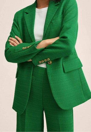 Femme portant un costume texturé vert vif avec un blazer et un pantalon assortis, les bras croisés sur un top blanc à col rond.